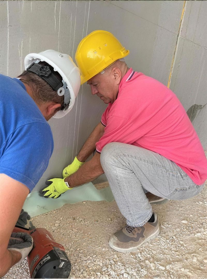Deux techniciens casqués posent une barrière anti-termites au pied d'un mur en construction